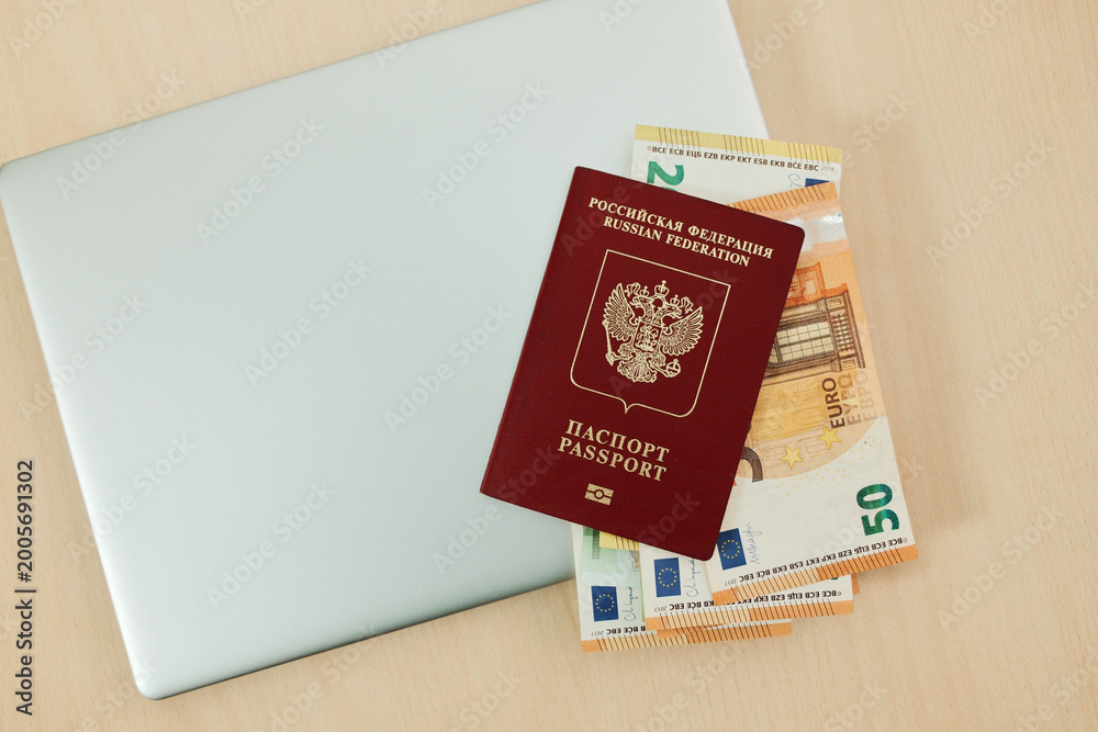 Fototapeta premium Laptop with Russian passport and 200 and 50 euro banknotes on wooden desk for immigration and travel planning concept. Online booking, relocation budget and digital vacation preparation.