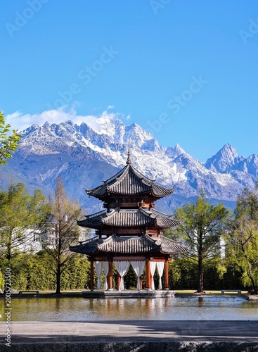 Traditional chinese wooden pavilion sitting beside a peaceful pond under high snowy mountain peaks during daytime