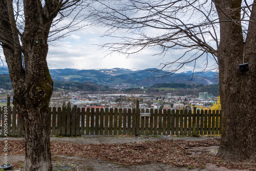 Obraz premium Baernbach, Austria – March 28, 2026: A panoramic view over a small town with houses, industrial buildings and distant mountains framed by trees and a wooden fence in the foreground.
