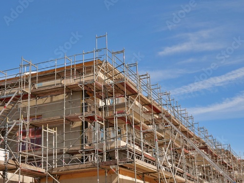 low-angle shot of a multi-story residential building completely enveloped in metal scaffolding during a major renovation or construction phase. The structure features wooden planks and safety rails un