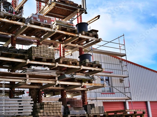 large industrial outdoor storage rack filled with various construction materials on wooden pallets. The rusty metal structure holds concrete blocks, paving stones, and buckets. In the background, a wh