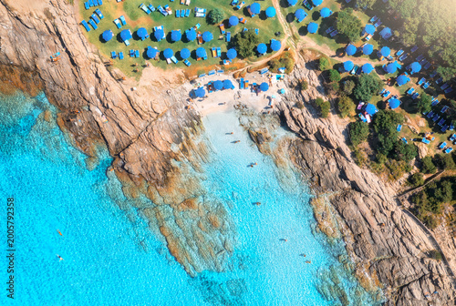 Aerial drone view of sea bay, sandy beach with umbrellas, stones and rocks in water at sunset in summer. Sardinia, Italy. Tropical scenery with sea lagoon, swimming people, blue water. Top down view