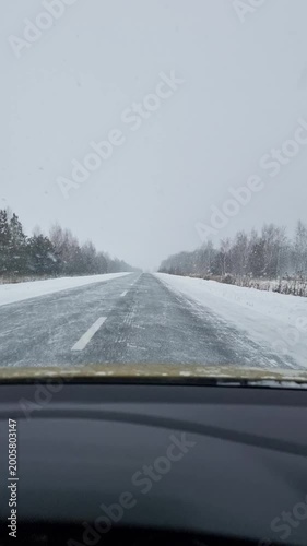 POV: driving on asphalt road during snowfall. Icy two-lane road during a blizzard: falling snow obscures the horizon and forest - hazardous driving conditions.