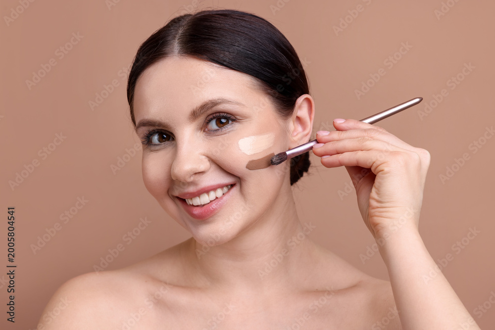 Fototapeta premium Woman applying makeup on her face with brush against beige background