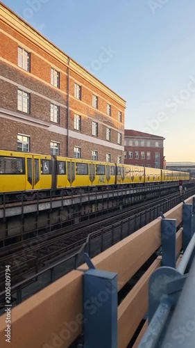 Subway train driving by on elevated tracks at sunset. Three quarter front view of a yellow U-bahn train driving along brick buildings during the golden hour.