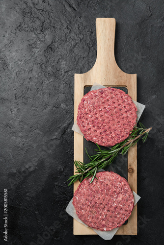Raw beef burger patties with rosemary on a wooden board, dark stone background with copy space, top view.