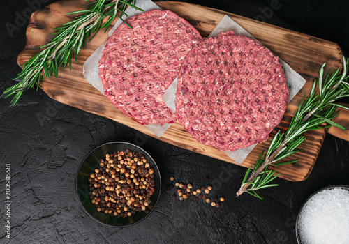 Close-up of raw beef burger patties on a wooden board with peppercorns, sea salt, and fresh rosemary, dark stone background, top view.