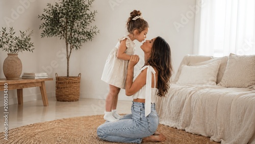Loving mother kneeling on a rug shares a tender kiss with her young daughter in a bright, minimalist living room filled with natural light and warm neutral decor.
