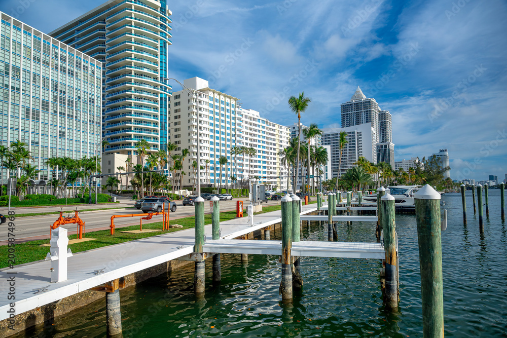 Naklejka premium Miami, USA, 16 April 2026: Waterfront pier overlooking coastal city buildings and road in Miami. Wooden pier with white posts next to high rise buildings and a street in Miami Beach Florida.