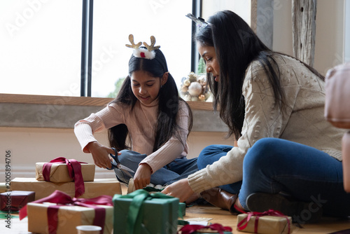 Indian mother and daughter wearing knit sweaters sitting at home wrapping kraft gifts with scissors