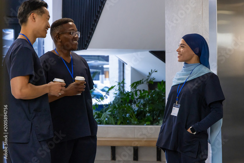 Diverse medical coworkers standing and talking in clinic lobby wearing navy scrubs holding coffee