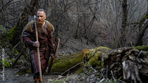 A rugged, bearded man dressed as a Viking stands confidently with an axe in a misty forest setting.