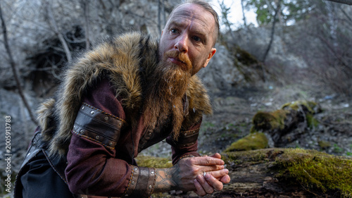 Φωτογραφία A rugged man with a long beard and fur-trimmed cloak kneels thoughtfully amidst a forest setting