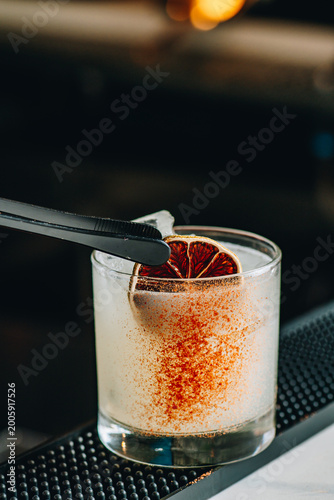 Craft cocktail served in a clear glass with a dried lime slice on a bar counter. A bartender prepares a craft cocktail with a slice of dried lime and spice.