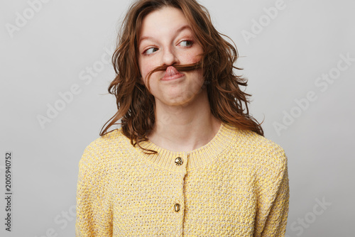 Fotografie Young woman with brown hair wearing yellow sweater makes mustache with hair, looking sideways with playful expression
