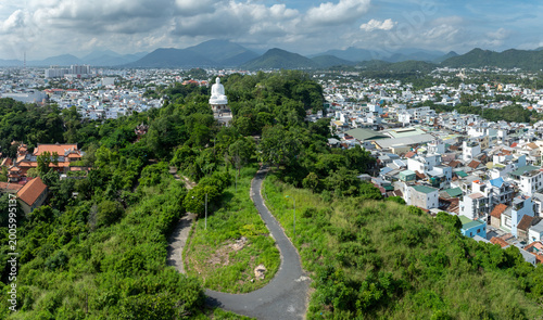 A panoramic view of the city of Nha Trang, Vietnam, with the iconic Lady Buddha statue overlooking the urban landscape and lush green hills at Long Son Pagoda.