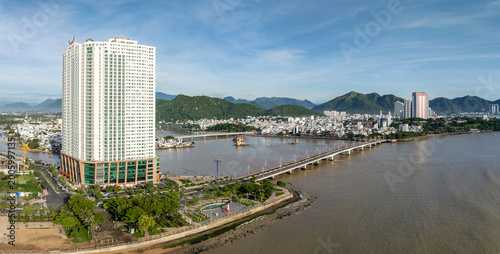 A panoramic view of the city of Nha Trang, Vietnam, showcasing the Cai River and the Bridge. The modern skyline is dotted with high-rise buildings and lush green mountains in the background.