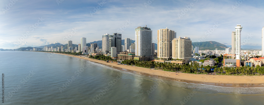 Fototapeta premium A panoramic view of Nha Trang beach in Vietnam, with modern high-rise hotels and buildings lining the shore. People enjoy the sandy beach and ocean waves under a partly cloudy sky.