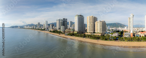 A panoramic view of Nha Trang beach in Vietnam, with modern high-rise hotels and buildings lining the shore. People enjoy the sandy beach and ocean waves under a partly cloudy sky.