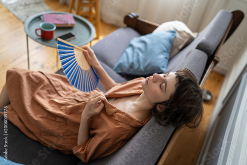 Billede på lærred Stressed woman suffering from heat at home sitting on sofa waving paper fan with closed eyes, female with dehydration and fever struggling with summer heatwave, trying to cool down with hand fan