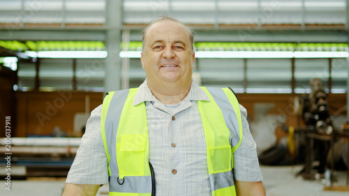 Cuadro en lienzo Senior Caucasian male factory worker wearing a yellow safety vest, smiling confidently while looking at the camera in a modern industrial warehouse background