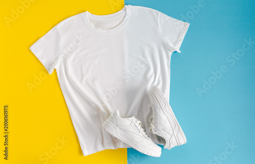 White blank t-shirt mockup with white sneakers arranged on a bright yellow and blue split background, top view.