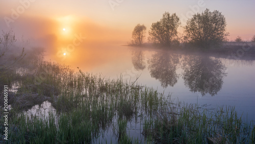 Fotografia A serene, misty sunrise over a calm river with glowing orange light and soft tree reflections
