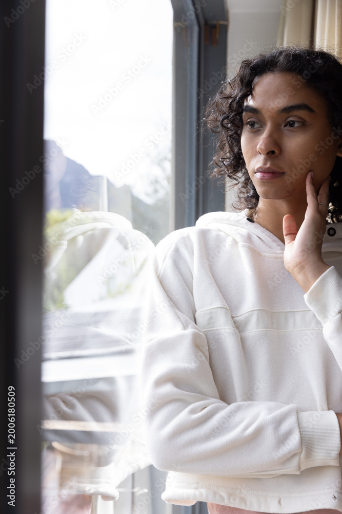 Fototapeta premium Adult nonbinary person standing at home by reflective glass looking out touching face in hoodie