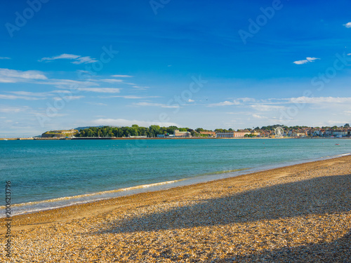 Panoramic view of a pebble beach overlooking a coastal town with Nothe Fort, a pavilion, and a funfair under a summer blue sky (Weymouth, England, United Kingdom)