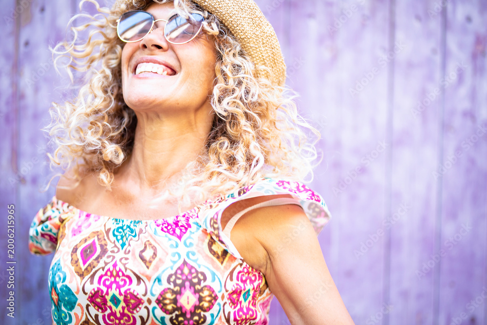Fototapeta premium Happy woman with curly blonde hair, sunglasses, and a straw hat is feeling joy and freedom while smiling and looking up against a lavender background, embracing a carefree summer lifestyle