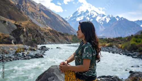 A woman stands by a river with mountains in the background