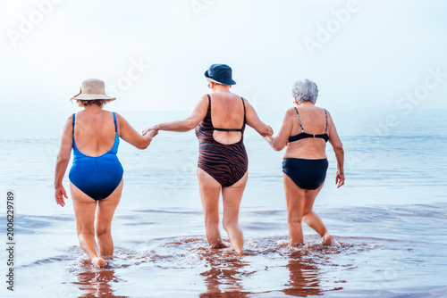Fotografie Three senior women friends holding hands, walking into the ocean water together,