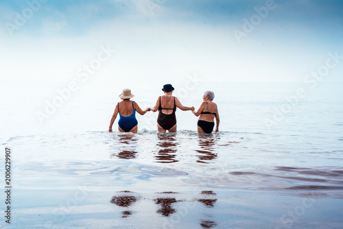 Three senior women wading into the ocean holding hands, sharing a moment of enduring friendship and active leisure during a summer vacation, reflecting companionship and wellness