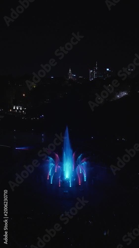 Vertical night drone footage captures Warsaw's magical glow over the Vistula. Park Fontann's lights dance on water, illuminating bridges and skyline in cinematic aerial sweeps