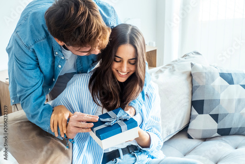 Obraz na plátne Man surprises woman with a blue gift box as she smiles and opens it on a cozy so