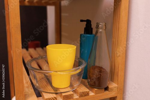 A yellow cup and glass bowl sit on a wooden rack next to bath products and a jar of brown substance