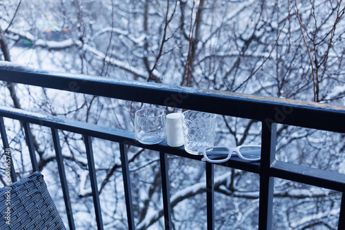 Glasses and a candle sit on a balcony railing with snow-covered trees in the background