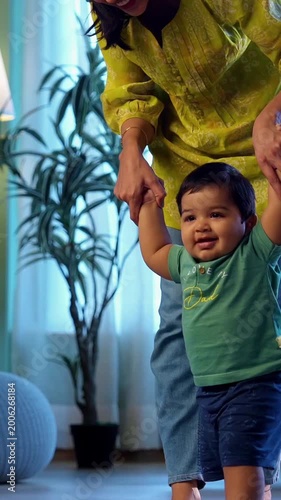 Indian infant boy walking holding young mother's hand in modern living room, learning to balance and take first steps with love, care, affection, and gentle guidance from mom