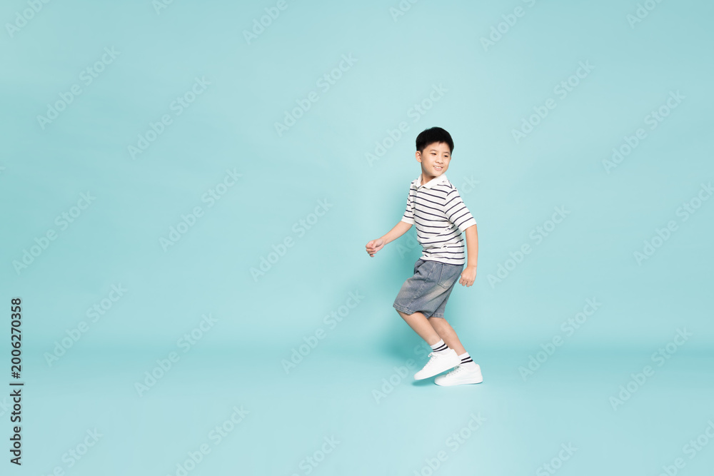 Fototapeta premium Happy young Asian boy in a striped polo shirt running cheerfully against a teal background. Childhood energy, active lifestyle, and healthy growth