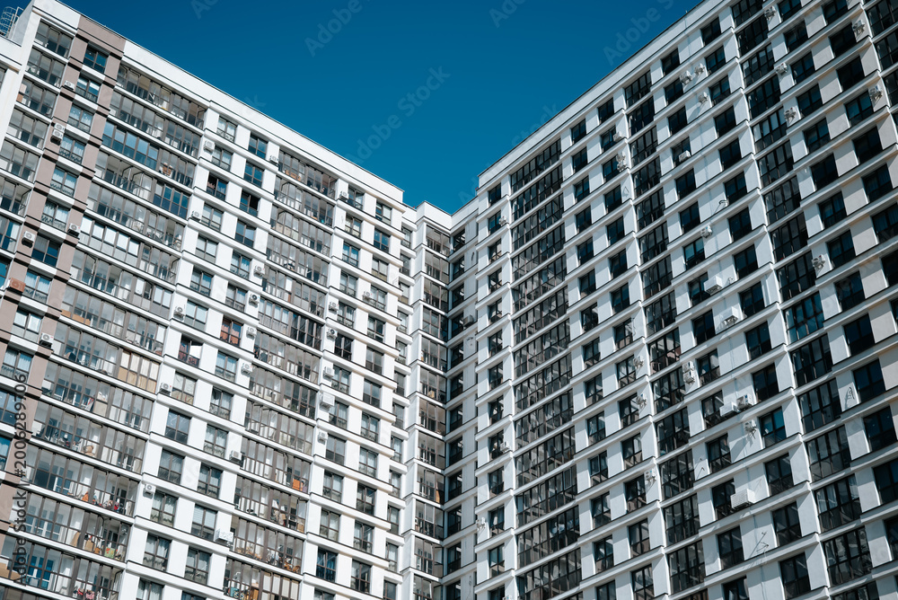 Fototapeta premium Two tall apartment buildings stand side by side under a clear blue sky. Their glass and concrete facades reflect the sunlight, showcasing urban living in a vibrant environment