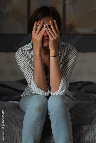 A young woman holds her hands over her face while seated on a bed in a softly lit room. The scene suggests feelings of sadness or distress as she appears lost in thought