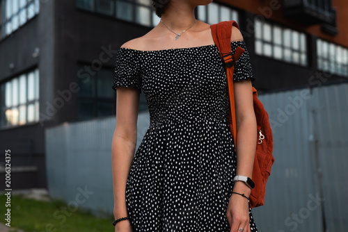 A woman stands in a polka dot dress next to an urban building in the evening light. She is holding a backpack and appears to be enjoying her time in the city