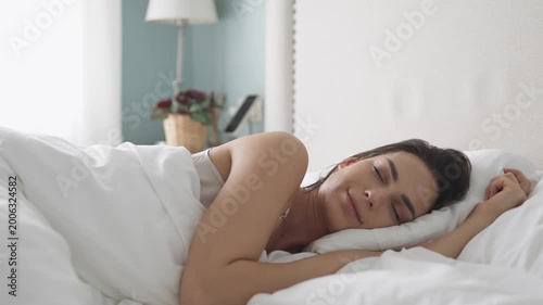 Woman waking up in a soft white bed, resting her head on a pillow, with gentle morning light illuminating the serene bedroom environment
