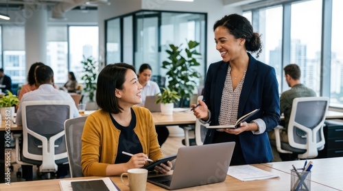 Two women are engaged in a collaborative discussion at a modern office desk. A senior businesswoman is guiding a younger colleague, who is working on a laptop.