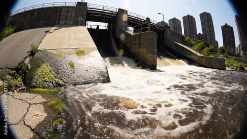 Low angle shot of water flowing over a dam with cityscape in the background on a sunny day