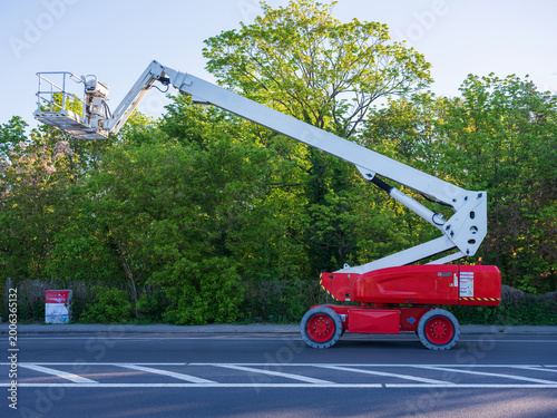 Картината върху платно Red and white articulating boom lift parked on a city street with green spring trees in the background
