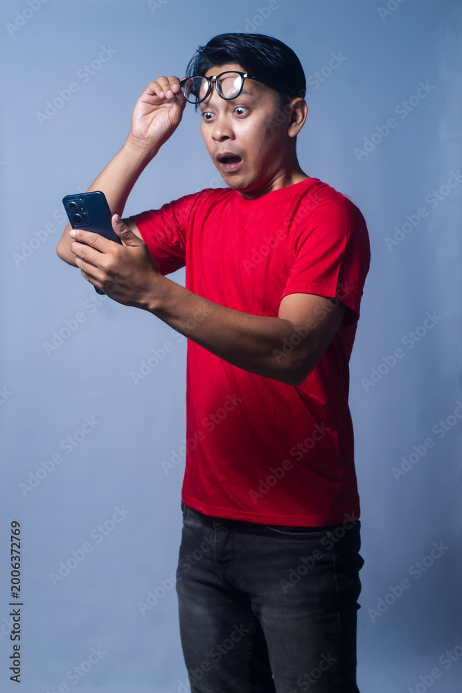 Fototapeta premium An expression of shock or disbelief is seen as an Asian man from Indonesia holds his cell phone and raises his glasses to his forehead. Isolated against a blue background.