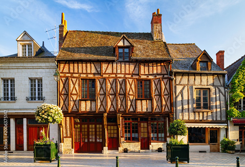 Sunlit historic half timbered houses with wooden beams, red doors, flower planters, and charming old town architecture under a blue sky, Amboise, France.