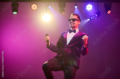 Singer presenter showman host performer with microphone. A confident man on stage under concert spotlight engages the crowd with upbeat energy. Concept of celebration, show, and entertainment.