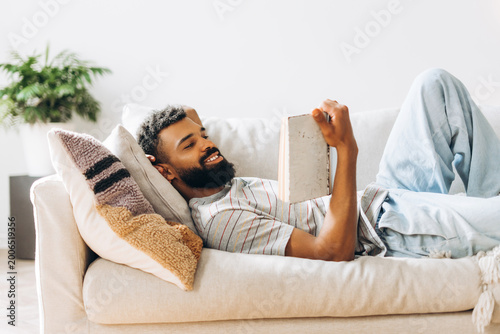 Obraz na plátně Young African American man lying on couch reading book at home